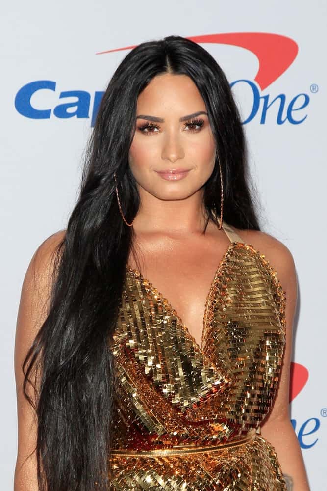 Demi Lovato wore a shiny golden dress to pair with her long layered raven hairstyle loose on her shoulders at the Jingle Ball 2017 at the Forum on December 2, 2017 in Inglewood, CA.