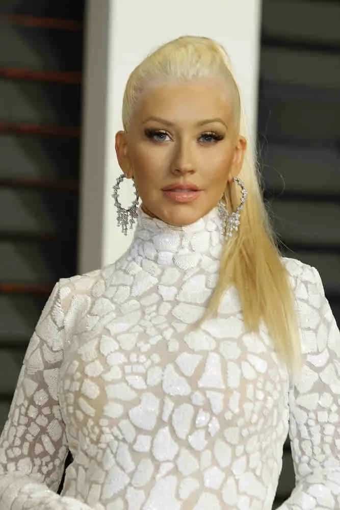 Christina Aguilera looks flawless in an all-white floor-length crack effects gown and her straight long blonde hair was styled into high ponytail at the Vanity Fair Oscar Party 2015 on February 22, 2015.
