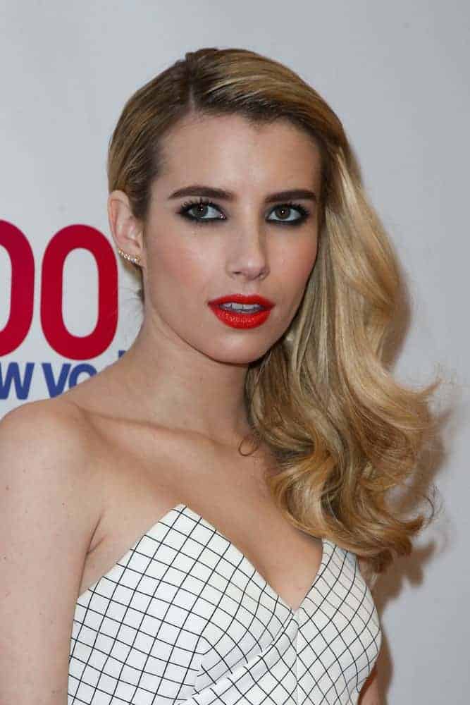 Actress Emma Roberts attended Z100's Jingle Ball 2014 at Madison Square Garden on December 12, 2014 in New York City. She wore a white patterned strapless outfit with her side-swept and highlighted wavy blonde hairstyle.