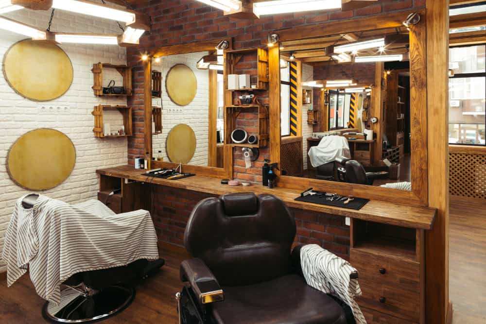 Here's an example of a small barber shop design with two chairs. As you can see it's fairly simple with white painted brick wall and then the hair cutting stations are against a dividing brick wall. Simple wood cabinets make up the stations. 