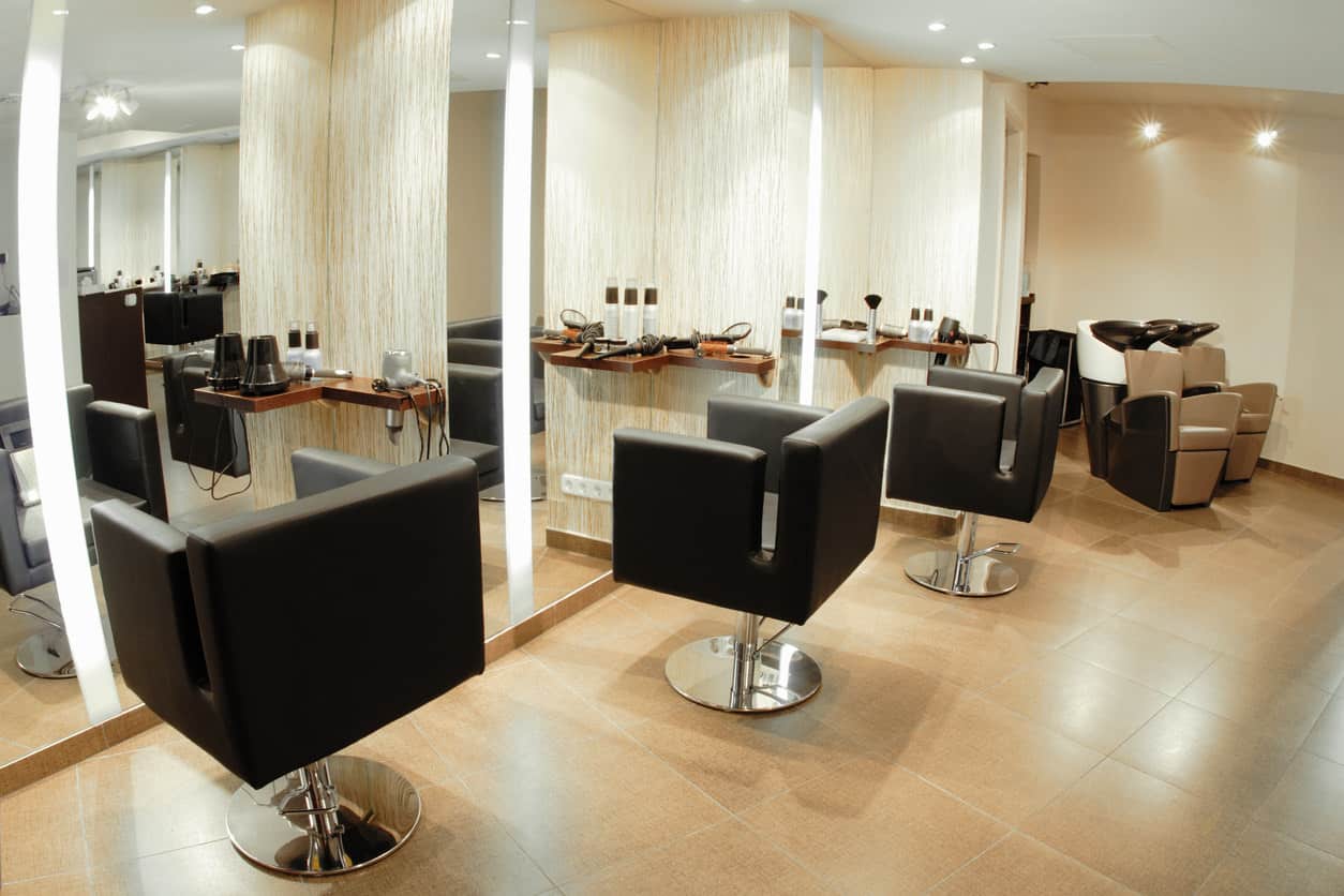This salon gives off a soft and warm ambiance with the cream colors and wood trim and tables. Paired with square-shaped black chairs with stainless steel bases, this salon has the feel of a 1950s barbershop. The textured wallpaper and flooring adds some interest and keeps it simple. The canister lights help to highlight certain parts of this salon and the white ceiling contrasts with the beige-colored walls and large tiled floor. 