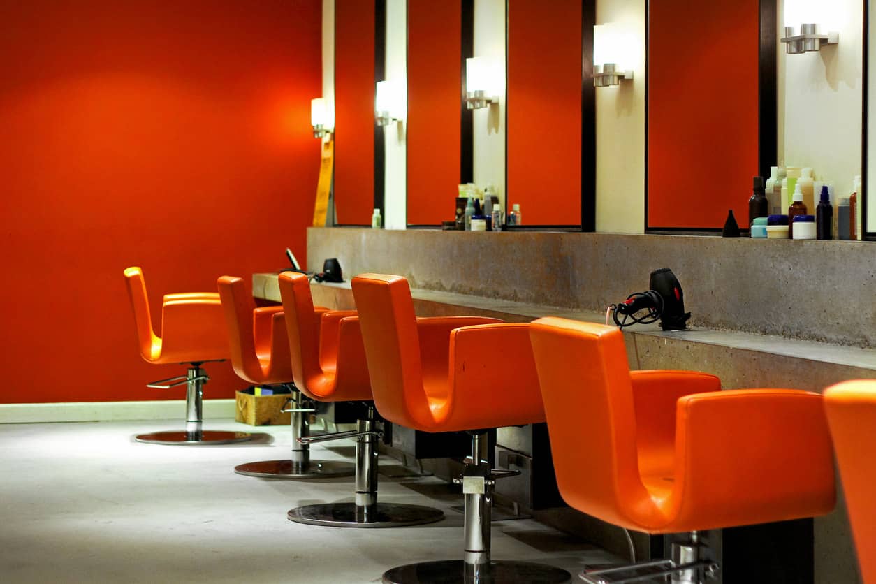 These bright orange chairs and walls of the same color paired with concrete tables and stainless steel light fixtures create a modern look. The concrete floors and black framed mirrors bring this modern look together. It has a warm feel although there is quite a bit of concrete used in this salon. 