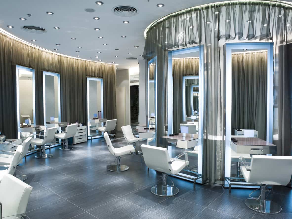 Countless adjustable flush mounted lights dot the ceiling to makes the salon feel like a stage where the customers are the stars. Sheer grey curtains hang from the ceiling, and oversized mirrors are in front of every stylist chair. The chairs are crisp white with see-through bases and armrests for a contemporary design. The mirrors glow with a baby blue light, and the light is reflected off of the semi-glossy grey textured floor. The bases of the chairs and the counters are stainless steel and reflect the light given off by the mirrors. 