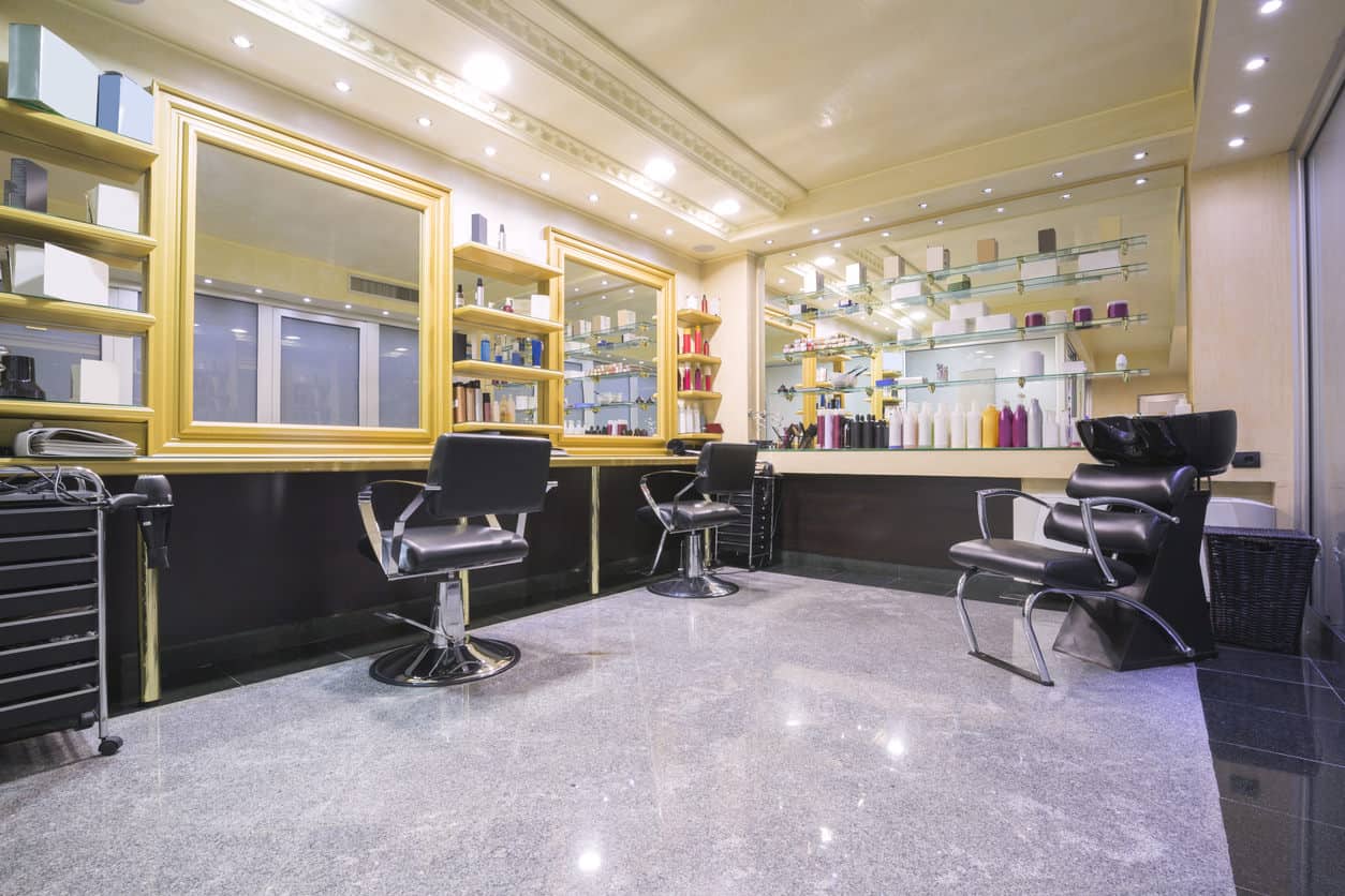 A raised ceiling accented by detailed trim adds some interest and opens up the room. Pale yellow walls and sunshine yellow frames around large mirrors and shelves brighten up this salon and offset the black chairs, dark tile, and dark wood. The glass shelves make them seem as if they are invisible and the colors of the products on them pop against the subtle yellow that's reflected by the mirror behind them. 