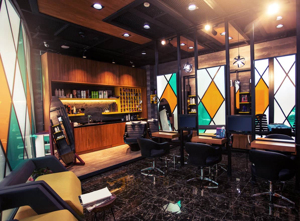 This salon features dark marbled tiled floor and rich wood cabinets and tables. Frosted white, yellow and teal diamond design of the stained glass offers a bold statement piece and compliment the wood nicely. The yellow and navy chairs and yellow shelves stand out against the dark marble and black countertop. This salon has the feel of a den or study, and you would expect customers to be lounging in their slippers and robe smoking a pipe. The metal globe pendant lights and an oval floor mirror are a nice touch. The black glossy marbled floor highlights the center of the room and adds to the richness of this salon. 