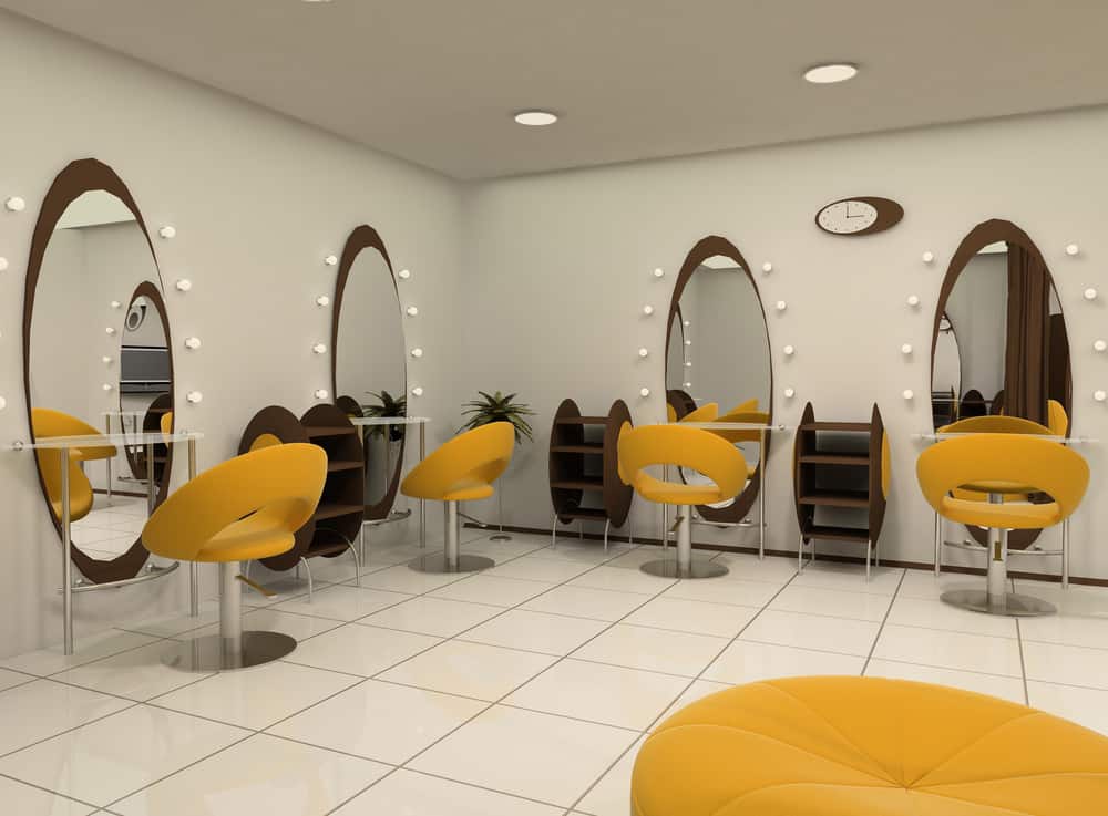 Futuristic yellow chairs and wood shelves stand out against the white walls and tile floor. Simple white bulbs frame each of the oval mirrors that are placed in front of the styling chairs. A bright yellow pouf looks bold against the large white tiled floor. 