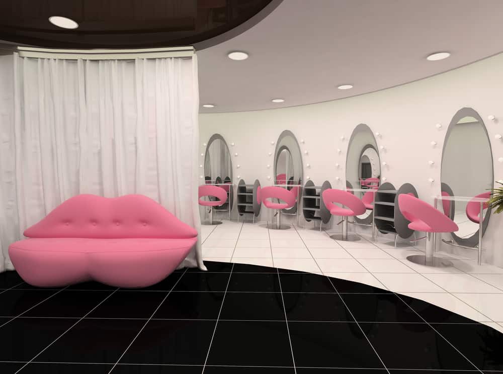 Fun bubblegum pink seating in the shape of lips greets customers when they visit this salon and the matching pink styling chairs really pop against the white walls and grey shelving. There is also a black tile accent on the floor that makes this salon very stylish and modern. The vanity lights around the oval mirrors stand out even when they are not lit. 