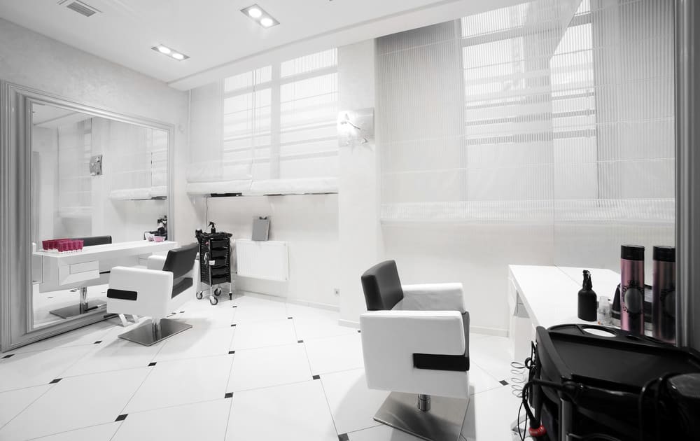 This almost entirely white salon has small accents such as the diamond pattern in the floor tiles and black sections of the stylist chairs. One wall is a mirror framed in a pale grey and the stainless steel base of the chairs add a hint of shine. 