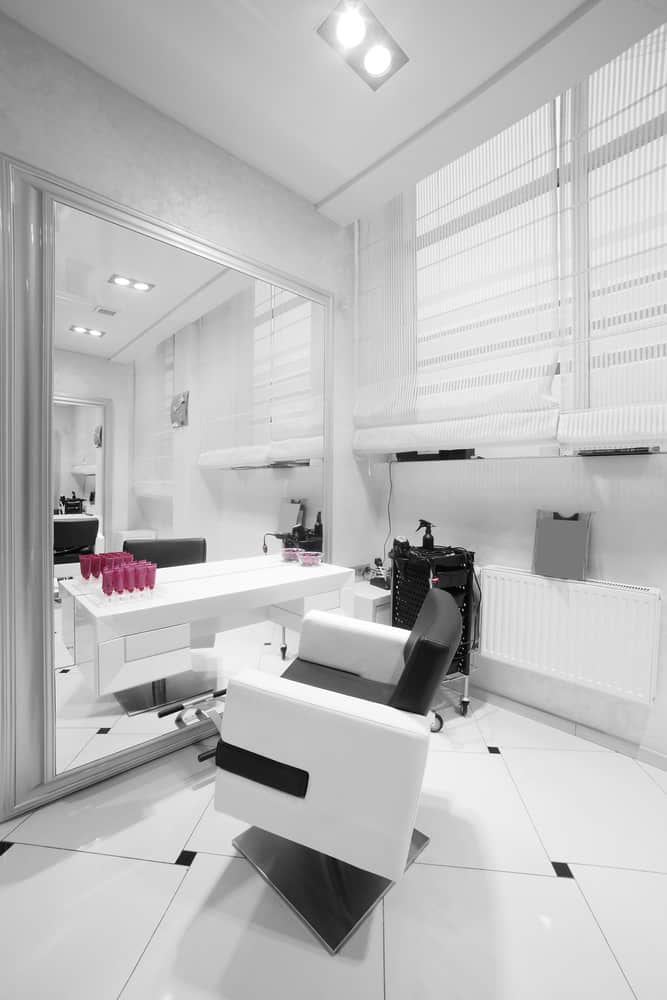 An almost all-white salon makes the rectangle tiles, portable cart, and pillow of the chair pop and draws the eye to the small details such as the blinds and the baseboard heater. 