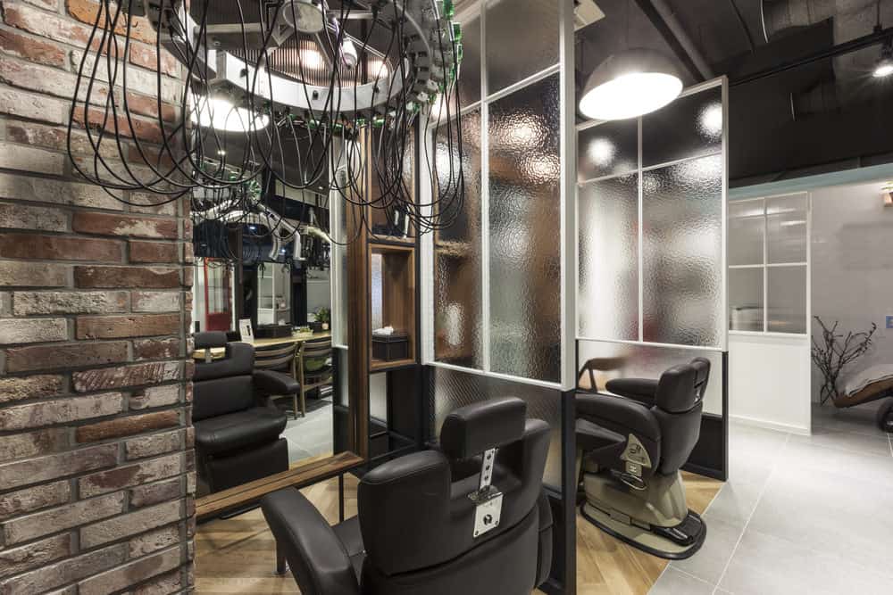 Interesting light fixtures immediately draws the eye upward, and the brick and textured window partitions create a relaxing and earthy feel. The soft black leather chairs and wood floor and accents inspire a calming atmosphere. A red door pops in the distance. 