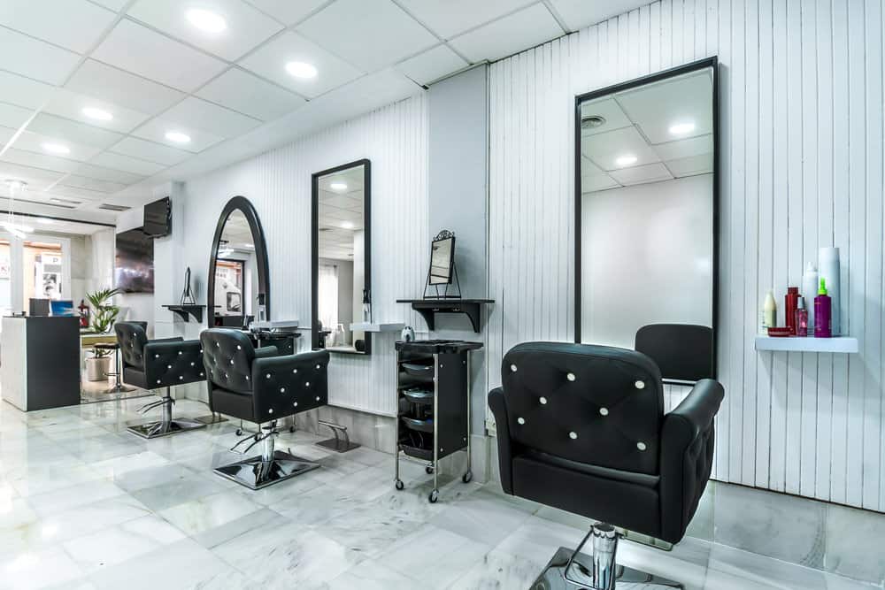 A white beadboard wall and black framed mirrors paired with the black and white buttoned chairs. The simple white shelves hold colorful products and the glossy marbled tile floors make this salon super chic and modern. 