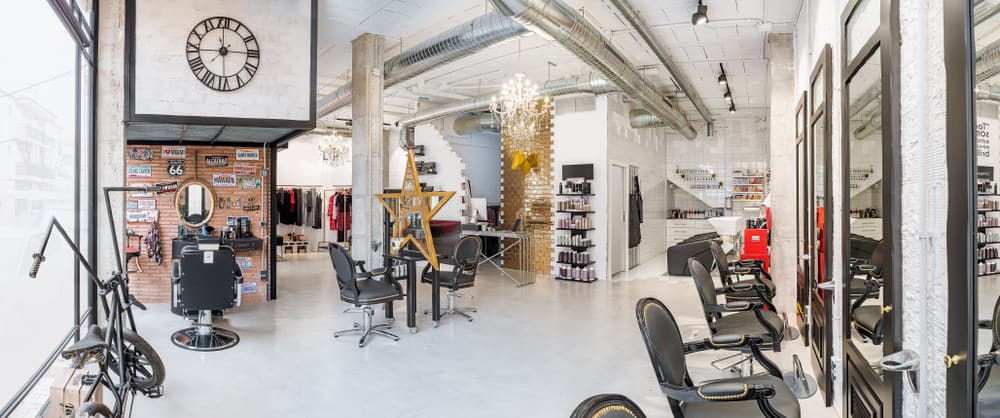This spacious warehouse has many focal points. The gold brick wall, golden star mirror, antique bike, license plate memorabilia displayed on the natural brick wall and the pops of red and yellow all stand out against the mainly white and grey background. The black leather and studded chairs look vintage, and the track lighting gives this salon an industrial look. 