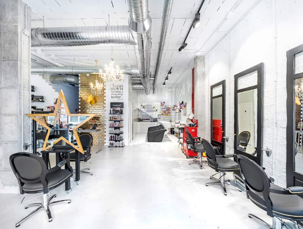The shiny metal ductwork is as much as a focal point as the gold brick wall and the red tool cart. The track lighting stands out against the white ceiling, and the warm glow of the chandeliers make this industrial salon look inviting. The black and stainless steel chairs look inviting, and the open space seems like it stretches on forever. 