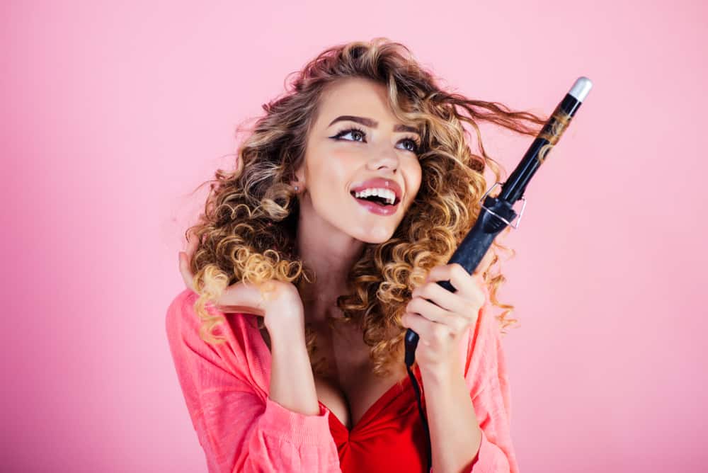 A woman with curly brown hair with a curling iron in her hand