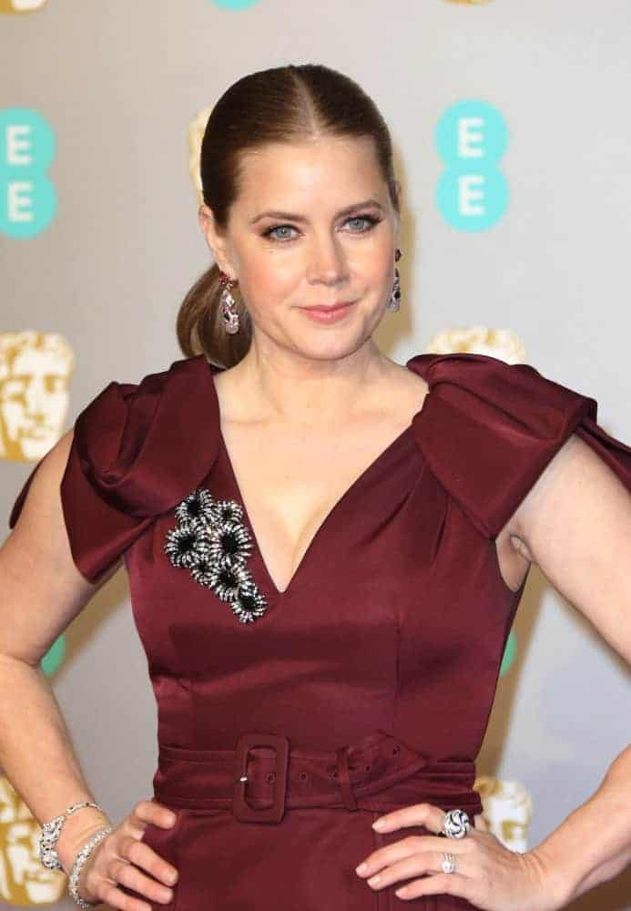 On February 10, 2019, Amy Adams attends the 72nd British Academy Film Awards at the Royal Albert Hall. She wore a maroon dress with a brooch to match her slicked-back auburn ponytail hairstyle.