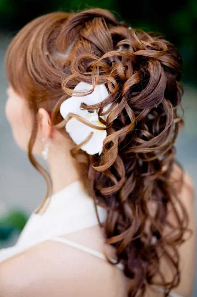 This hairstyle for beautiful long hair consists of a ponytail-style hairdo with several lacy strands. A pristine white flower adds grace to the hazelnut shade of hair.