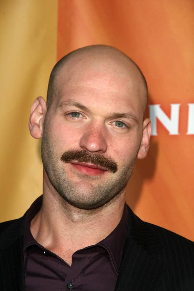 Corey Stoll has never looked better than in this classic clean head, except with slight hair on the sides coupled with a prominent chevron mustache! The icy blue-green eyes with shades of grey and that slightly darker facial scruff take the entire look to a whole new level.