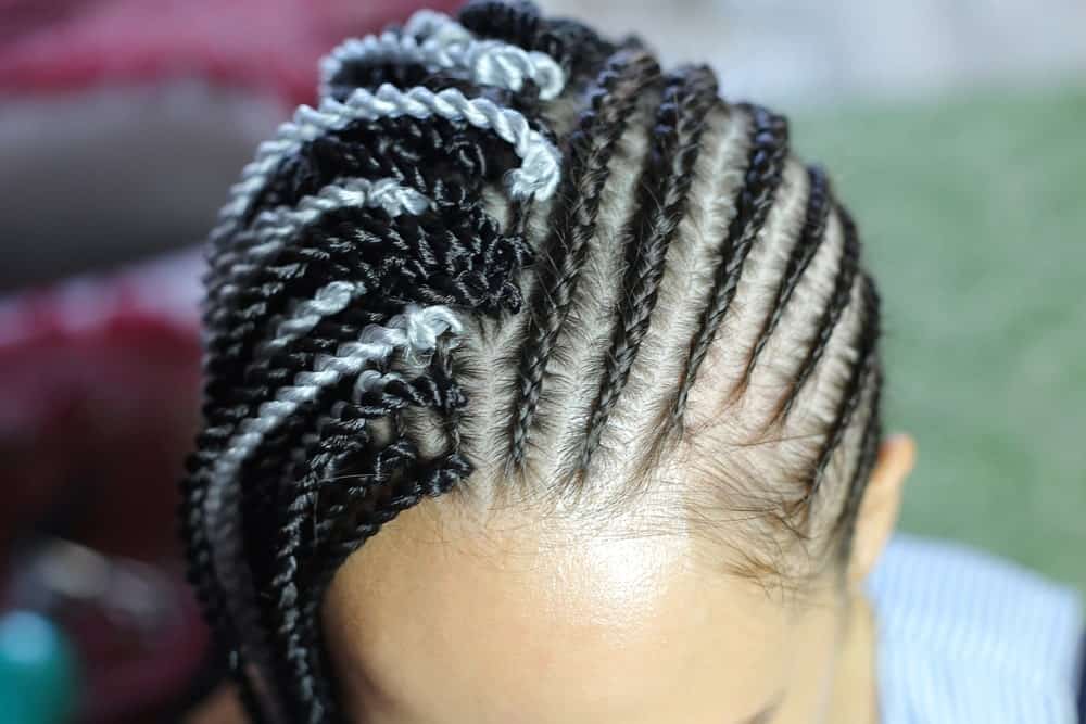 Why only style your Fulani braids one way when you can style them in two different ways! Simply part your hair in the middle and use one side to create braids that are running from the front to the back and the other side to weave braids that are running to the side and down. To add some extra oomph to the style, add some white yarn thread to your dark hair for a very cool and urban monochromatic look.