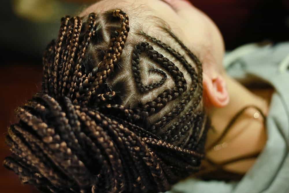 Fulani braids are extremely creative and versatile and incorporate innumerable elaborate tribal patterns. This model is sporting a geometric, maze-like pattern, formed from braids, on the sides of her head. To give some more dimension to her hair, she has added antique gold-colored yarn.  You can keep your braids down for this style or wrap it up in a ballerina bun like this model.