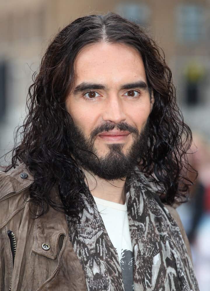 There is probably no one who rocks long curly hair better than Russell Brand! He has really long, curly black hair that falls on both sides of his face through a clean middle parting. The hair seems to have been gelled from the front since it is giving off a very wet, shiny look. The addition of the black beard and mustache further enhance the overall look and give him a very mature, macho look.