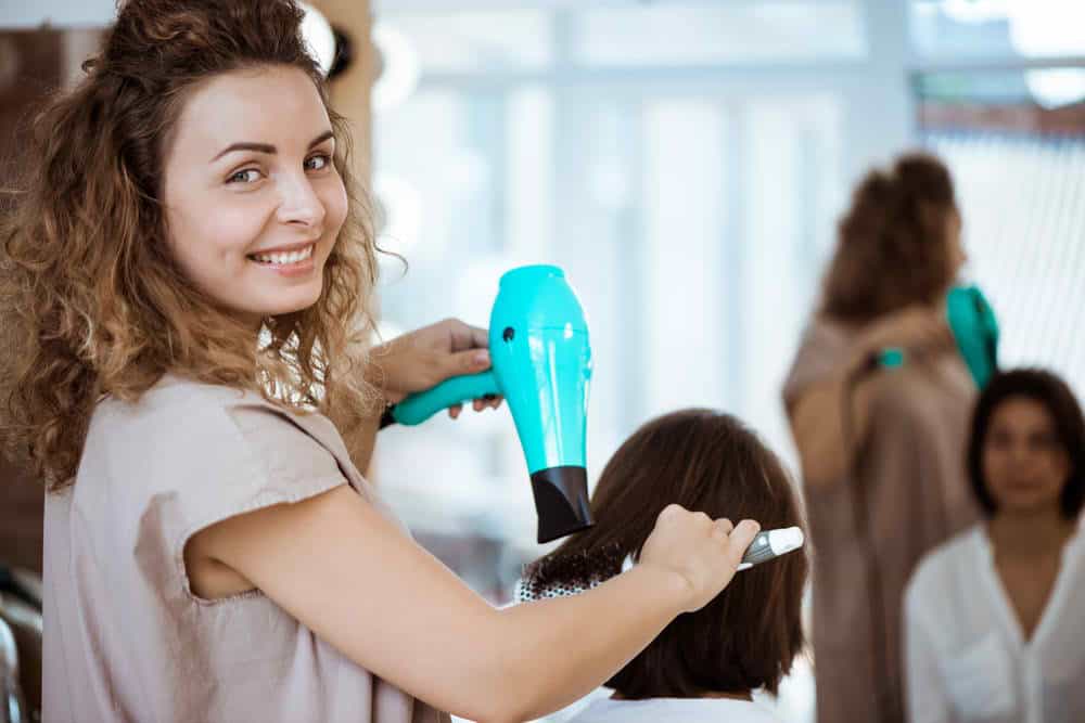 A hairdressser looks into the camera with a smile while hairblowing a customer's hair.