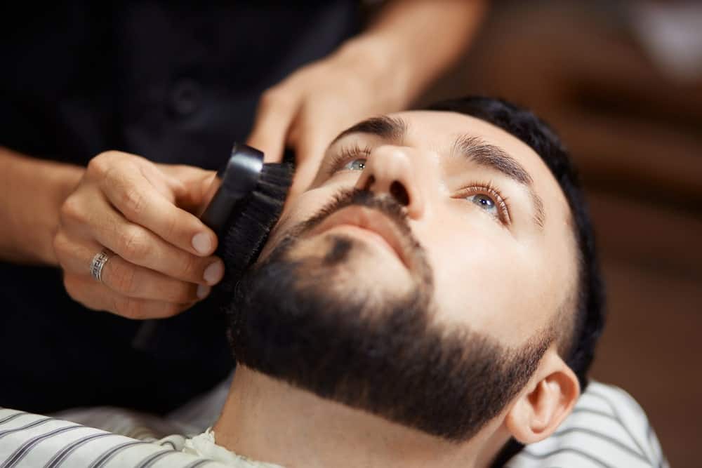 Man lying on a barber shop chair while the barber brushes his well-maintained beard.