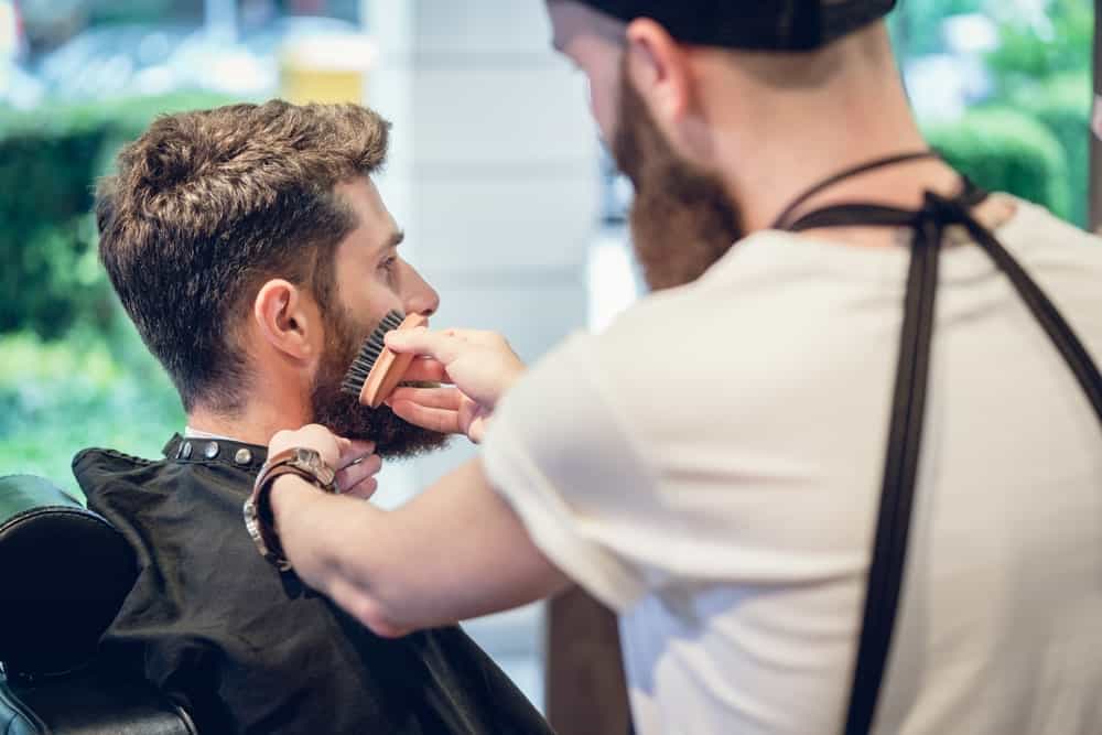 Man in a barber shop with a nice haircut and a well-maintained beard. His barber is brushing his facial hair.