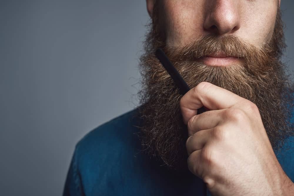 Man with thick beard in a blue shirt, combing his beard.