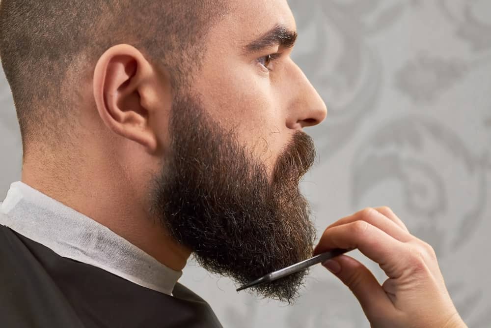 A side view of a man with thick beard in a barber shop, combing his beard.