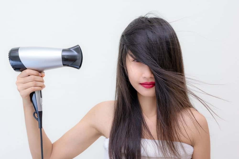 Heat Gun Vs Hair Dryer. A woman using a hair dryer on her long dark hair.