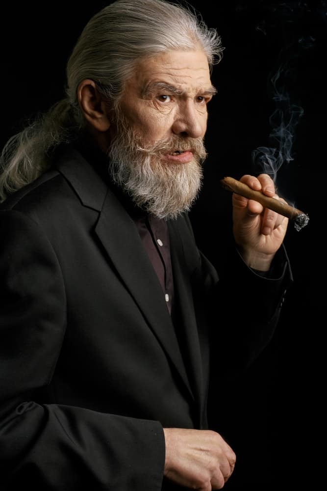 A white-haired old man rocks a ponytail and beard while smoking cigar.