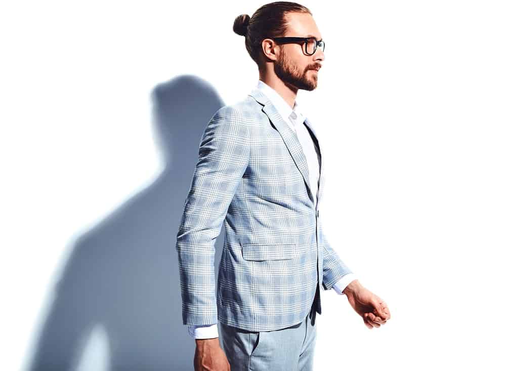 A look at a young businessman in a plaid blazer covering the white inner shirt on a white background with his shadow showing up. He has a beard and a man bun hairstyle, along with an eyeglasses.