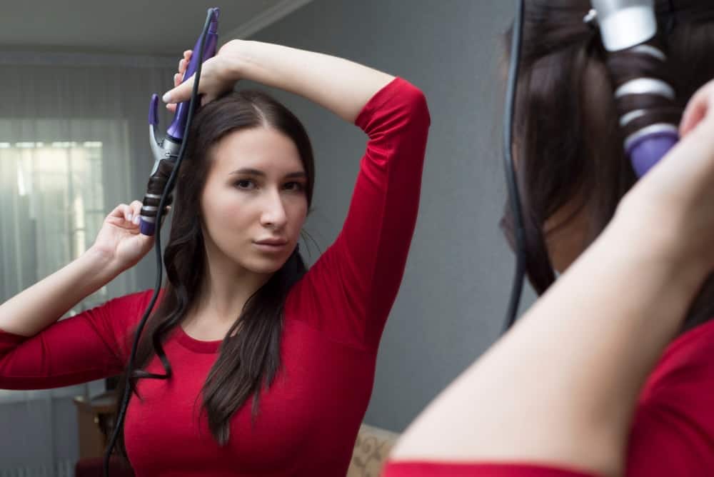 A brunette woman using a heated hair roller in her room.