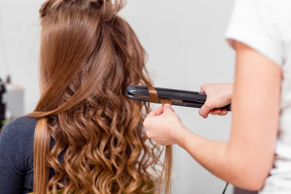 Back view of a woman's hair being curled by a hairdresser using a curling iron.