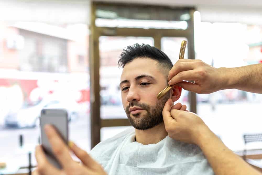 A man checks his phone while getting a haircut.