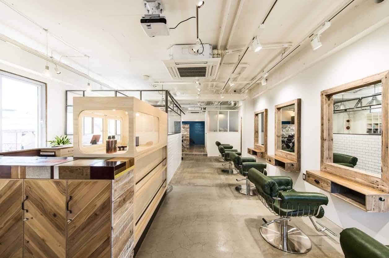 Hair salon interior with white walls, concrete flooring, chevron reception, and green leather chairs paired with wooden framed mirrors.