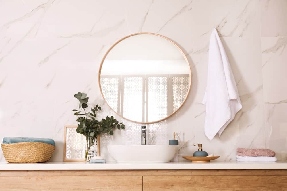 Wooden vanity with vessel sink and a round mirror fixed against the white marble wall.