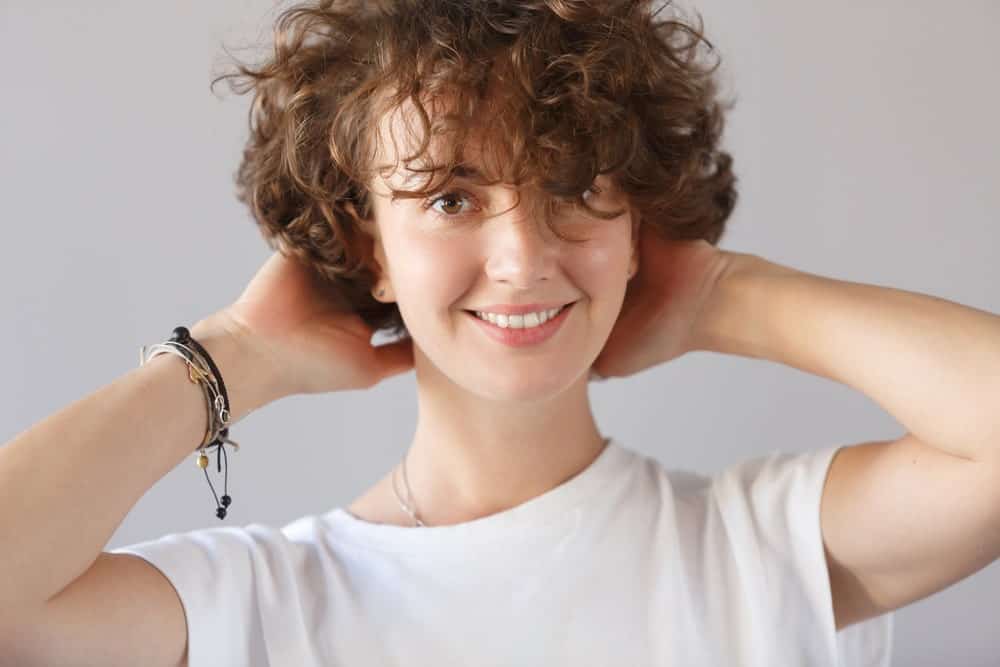Woman with short curly hair and bangs holding the back of her head.