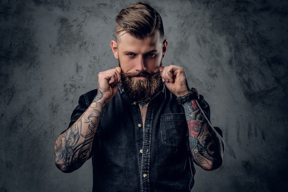 Man with sleeve tattoos styling his beard.