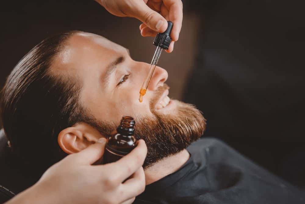 A barber applying oil on a man's beard through a dropper.