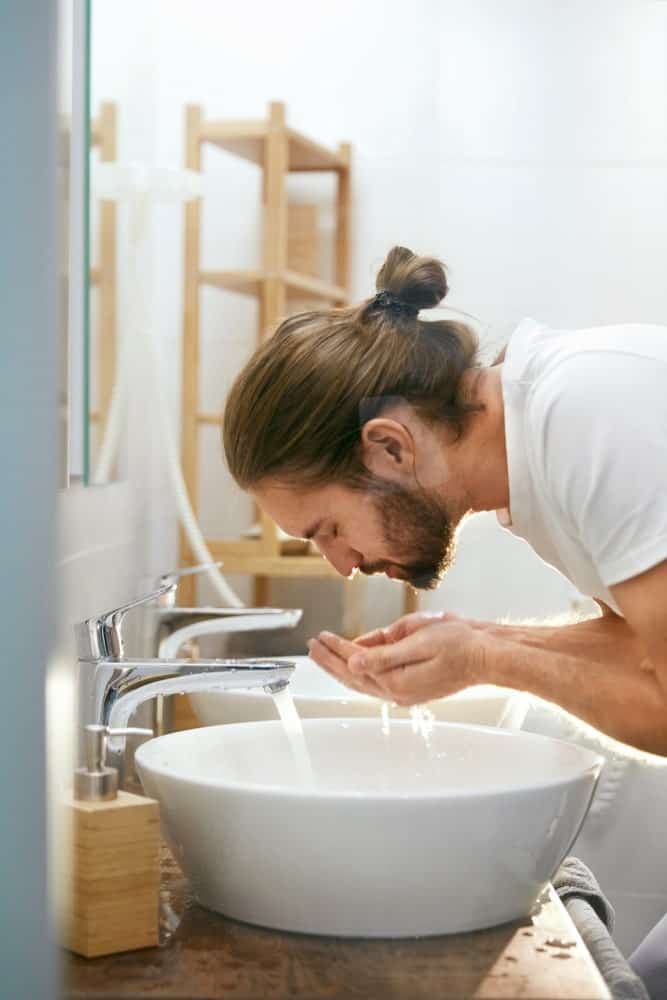 Man with full beard washing his face on the bathroom sink.