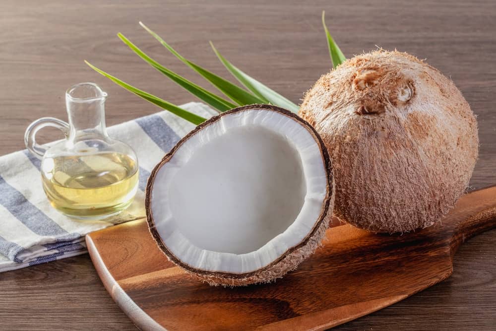 A glass bottle of coconut oil next to raw coconuts.