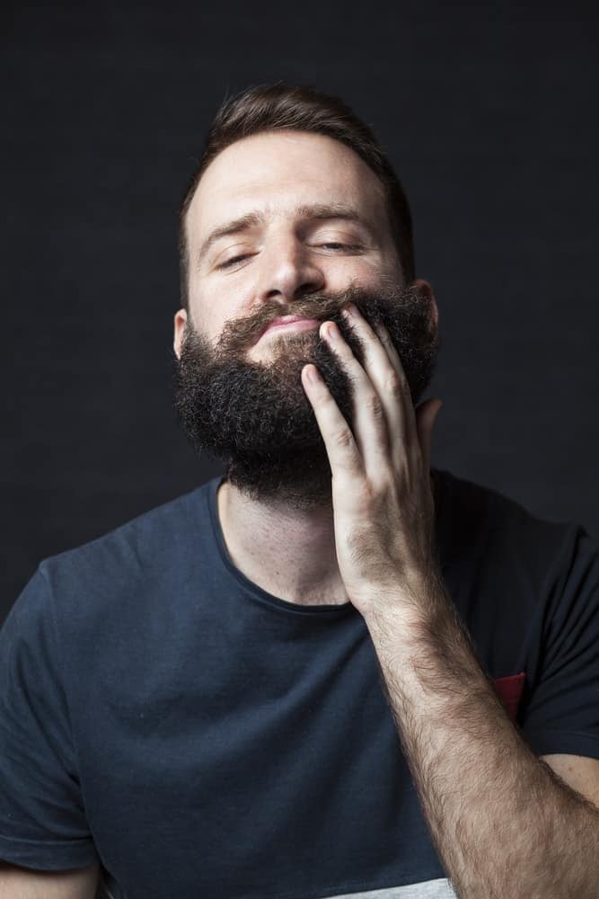 A bearded man applying beard oil on his thick beard.