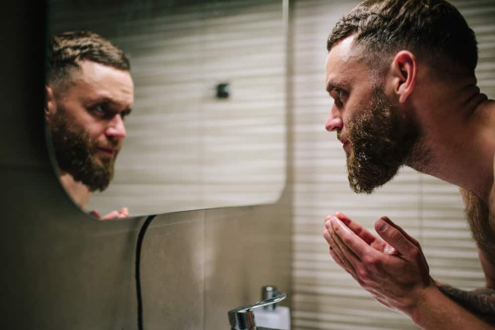 A bearded man washing his face at the bathroom sink.